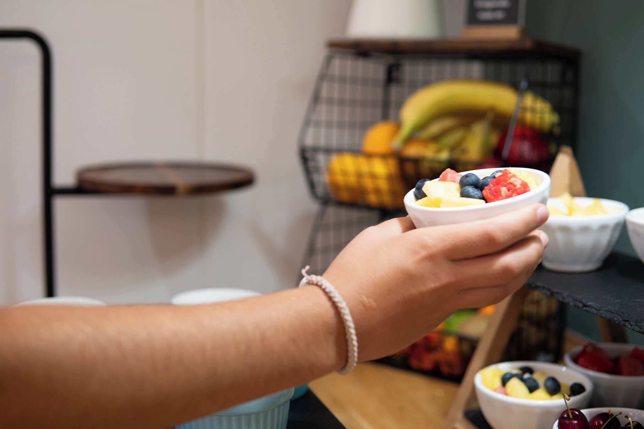 Eine Hand hält eine Schale mit Obstsalat, im Hintergrund ein Korb mit Bananen und anderem Obst.
