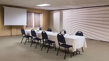 Un salón de conferencias con una larga mesa, sillas, una pantalla de proyector y una ventana con persianas.