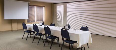 A conference room with a long table, chairs, a projector screen, and a window with blinds.