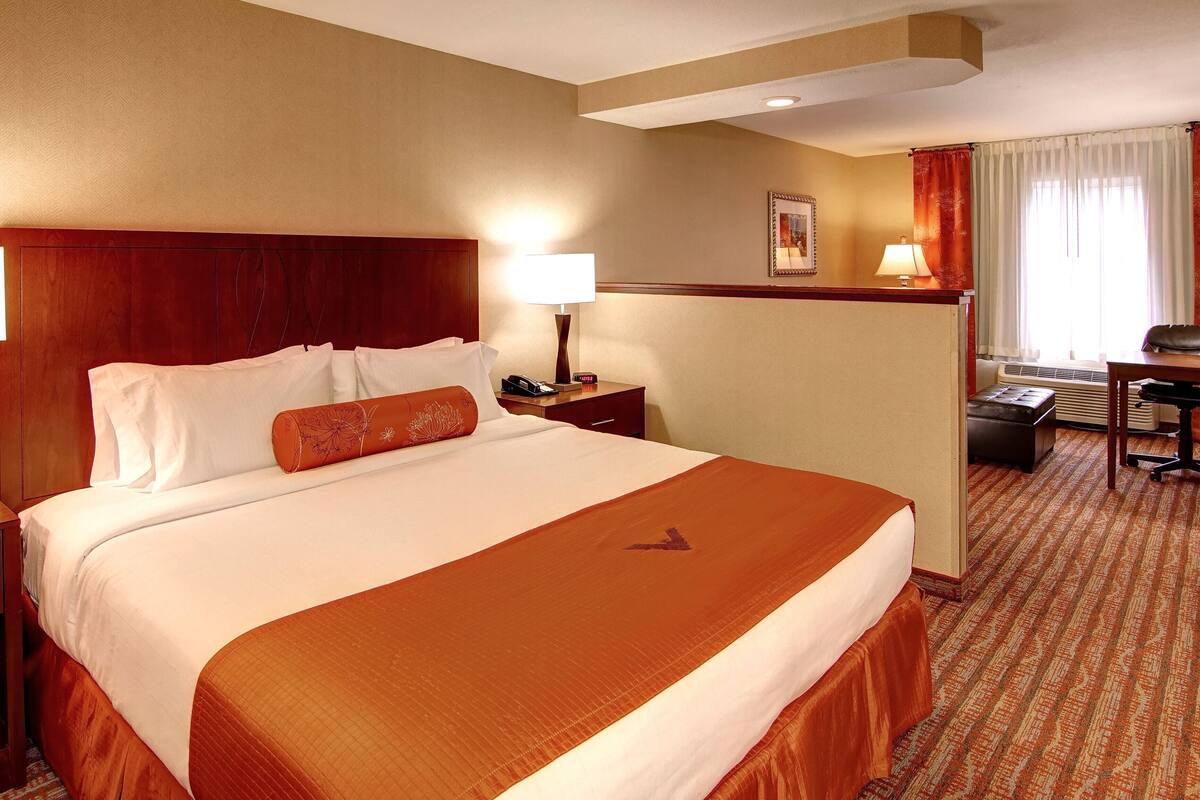 Quarto de hotel com uma cama grande, uma escrivaninha, uma cadeira, uma mesa de cabeceira e um abajur.