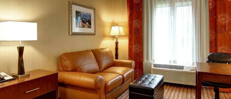 A hotel room with a leather sofa, a small table, a lamp, a picture on the wall, and a window with curtains.