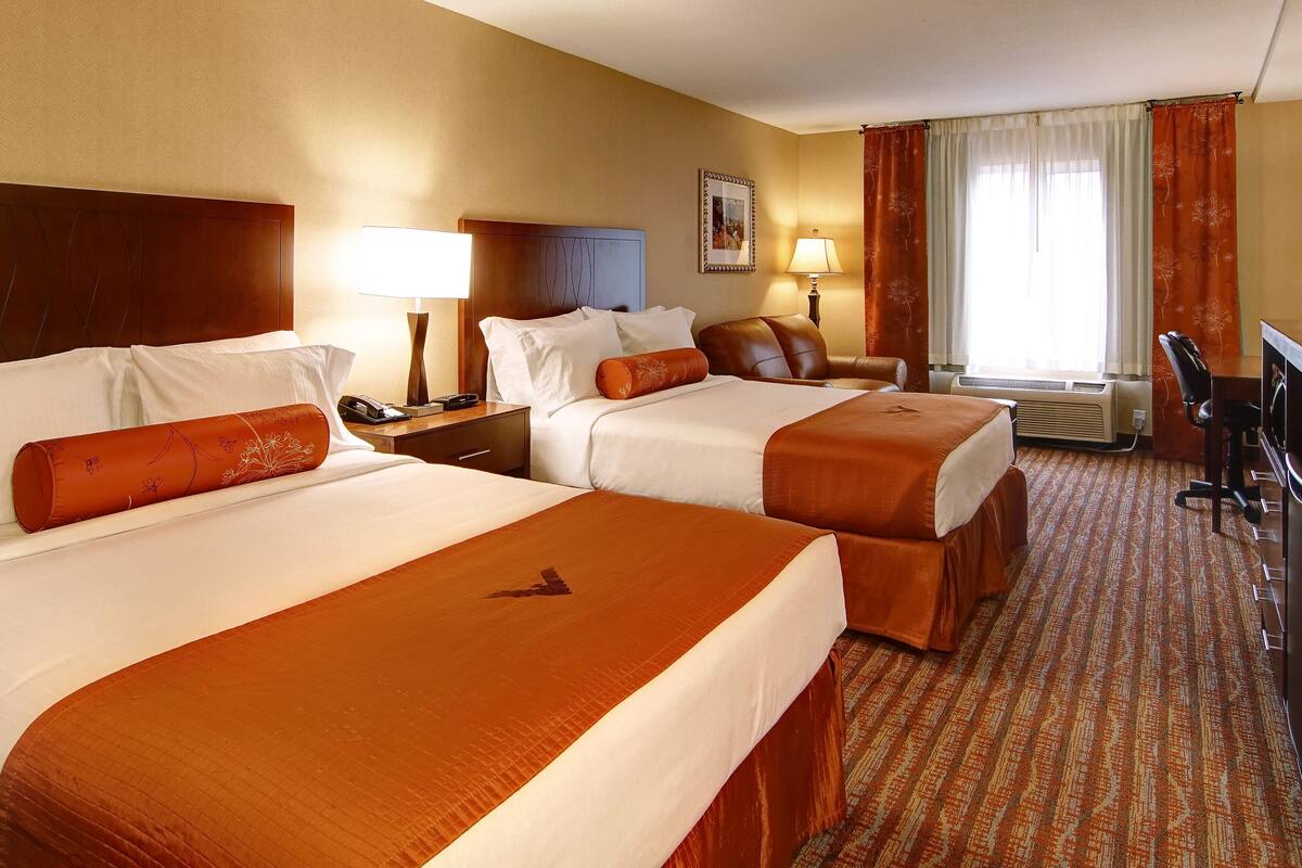 Quarto de hotel com duas camas, uma escrivaninha, uma cadeira, um abajur e uma janela com cortinas.