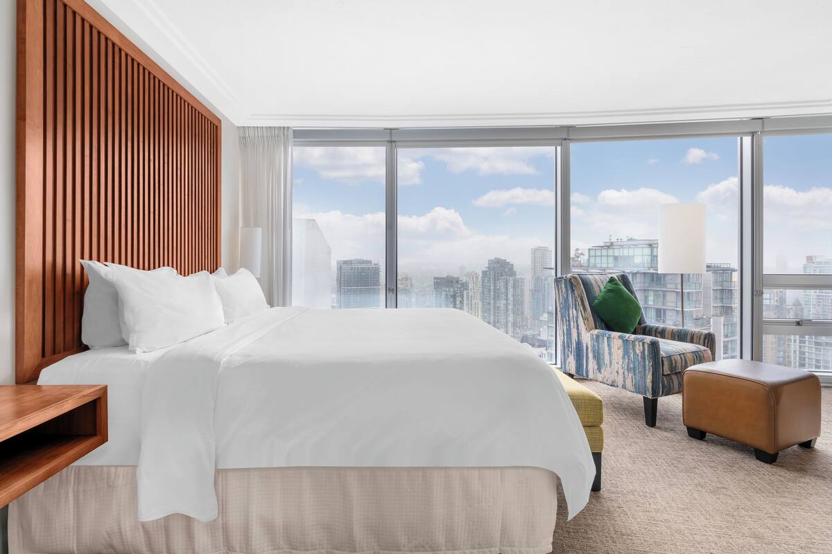 Een hotelkamer met een groot bed, een houten hoofdbord, uitzicht over de stad, een bank en een poef.