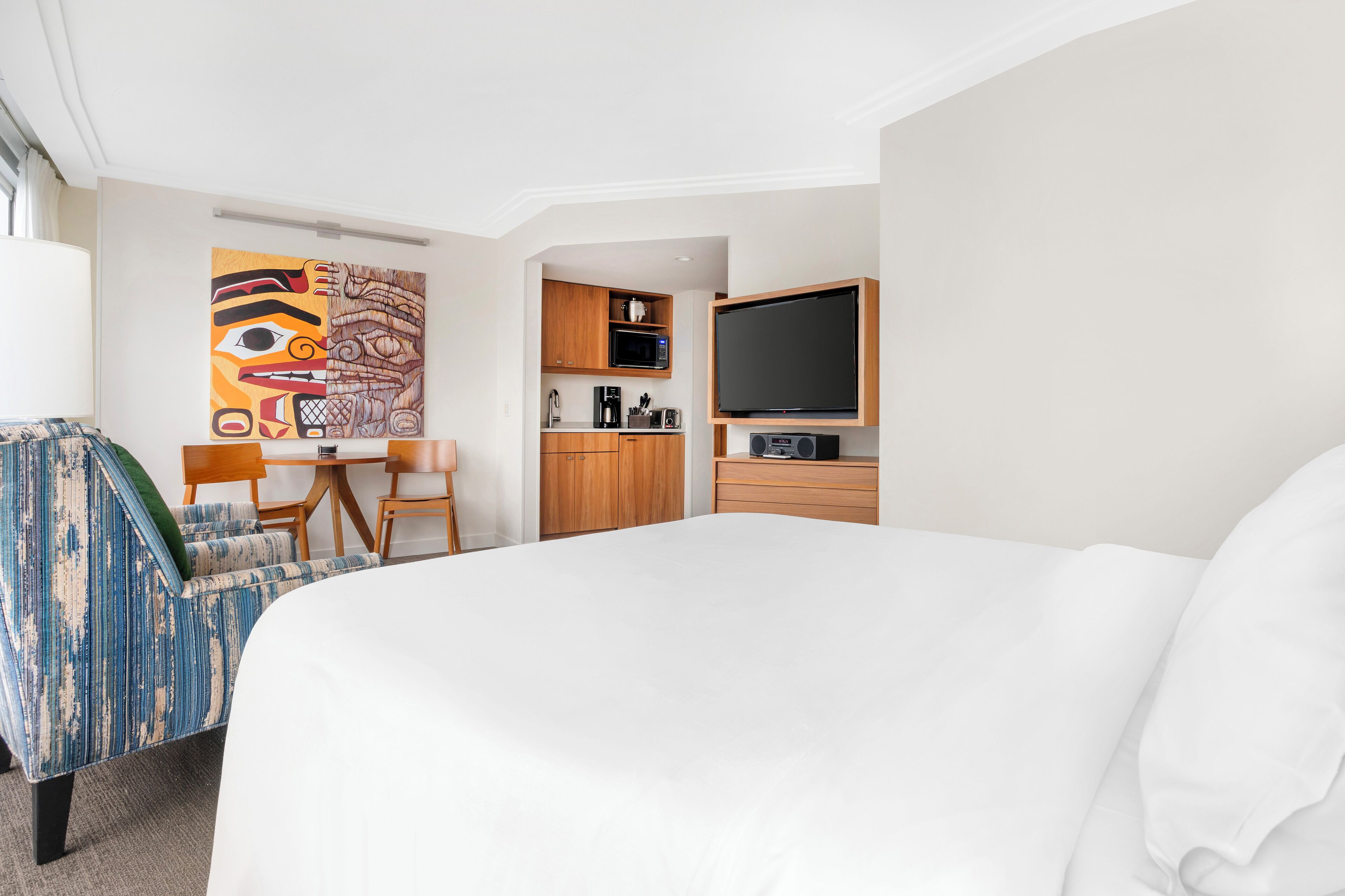 A hotel room with a bed, a desk, a chair, a microwave, a coffee maker, and a flat-screen TV.