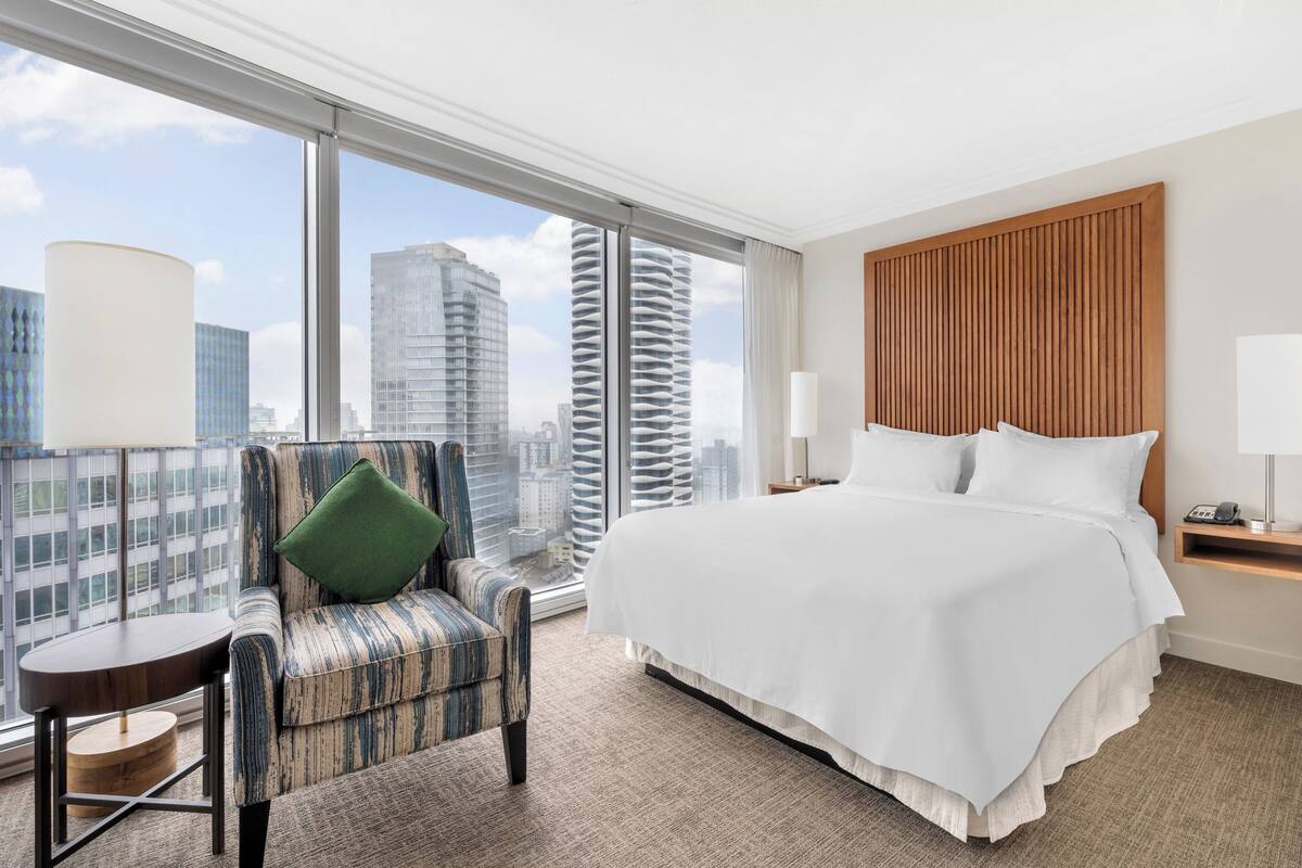 Een hotelkamer met een groot bed, een stoel, een bijzettafeltje en uitzicht op de stad door grote ramen.