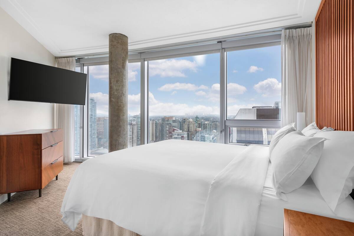 Hotelkamer met een groot bed, een flatscreen televisie, een houten dressoir en uitzicht op de stad door grote ramen.