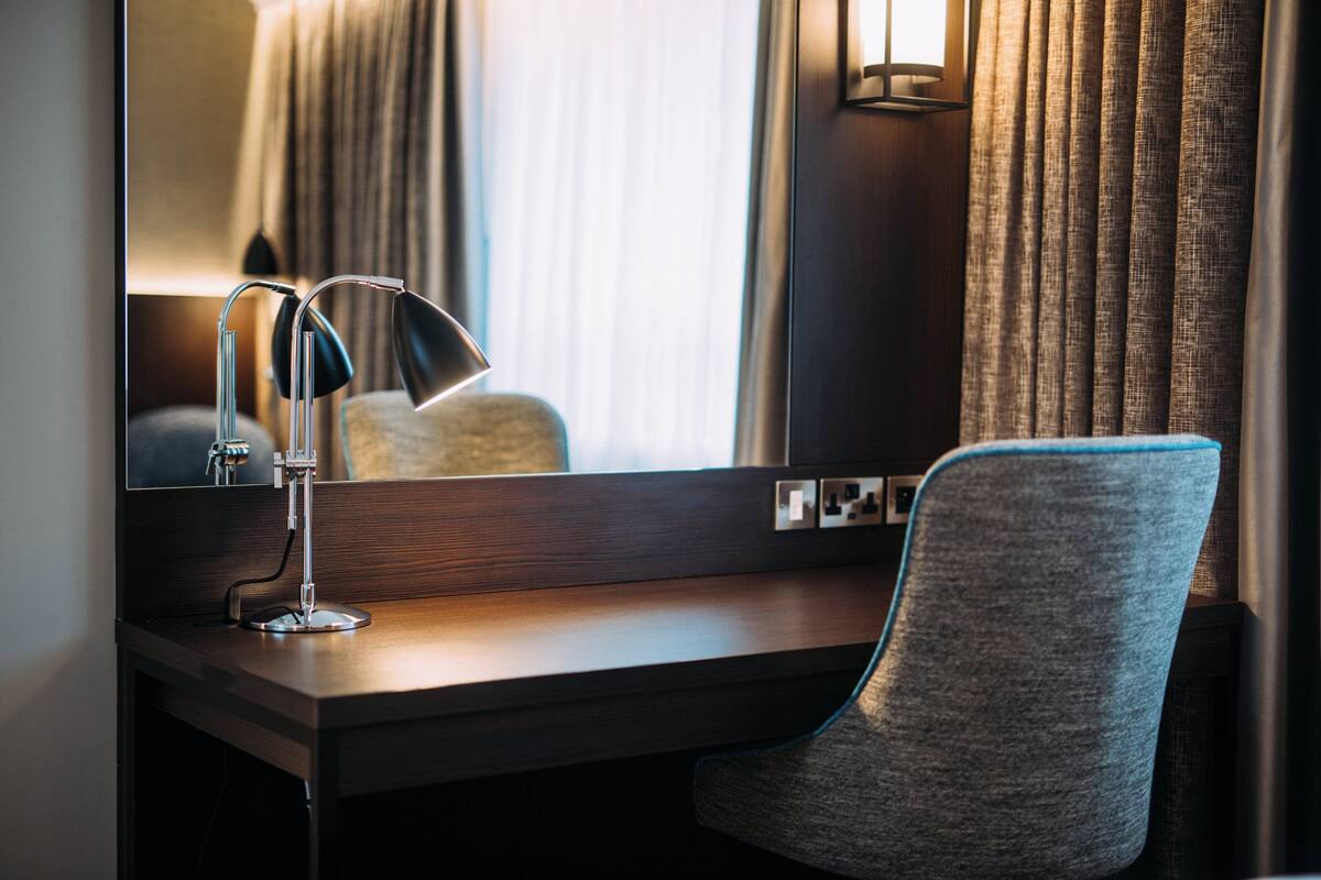 A hotel room with a desk, a chair, a lamp, a mirror, and curtains.