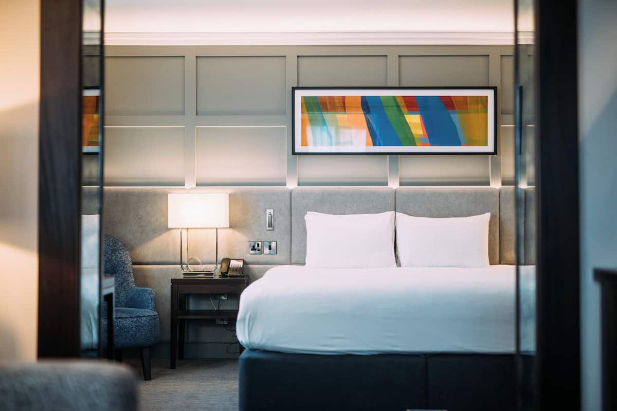 A hotel room with a bed, a chair, a nightstand, a lamp, and a colorful abstract painting on the wall.