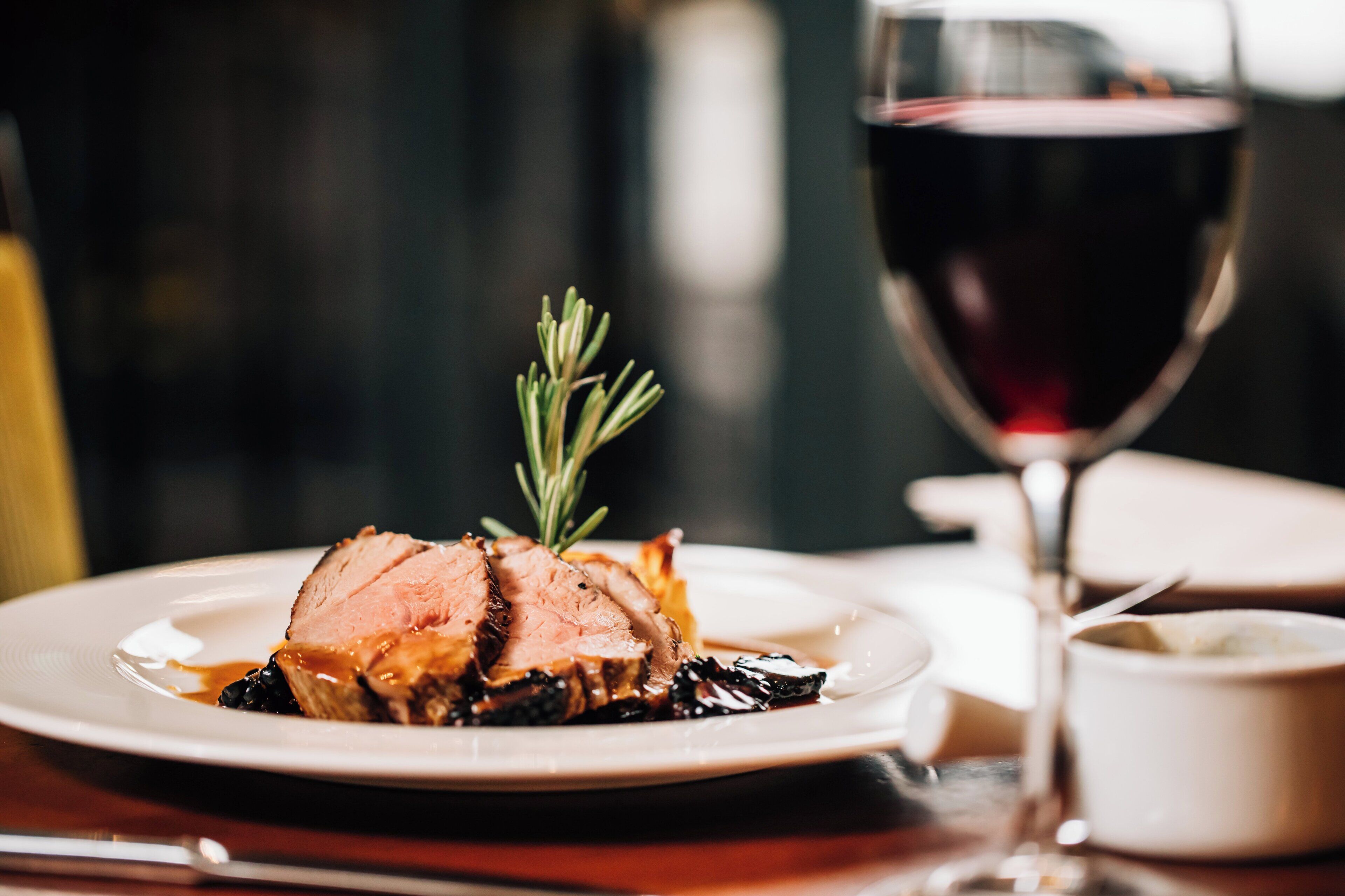 Une assiette de viande tranchée, garnie d’une branche de romarin, accompagnée d’un verre de vin rouge et d’un petit bol de sauce, le tout posé sur une table en bois.