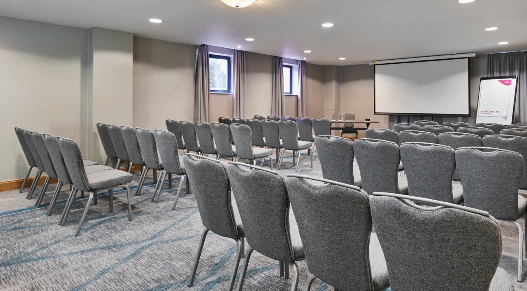 Une salle de conférence avec des rangées de chaises grises, un écran de projection et un tableau blanc.