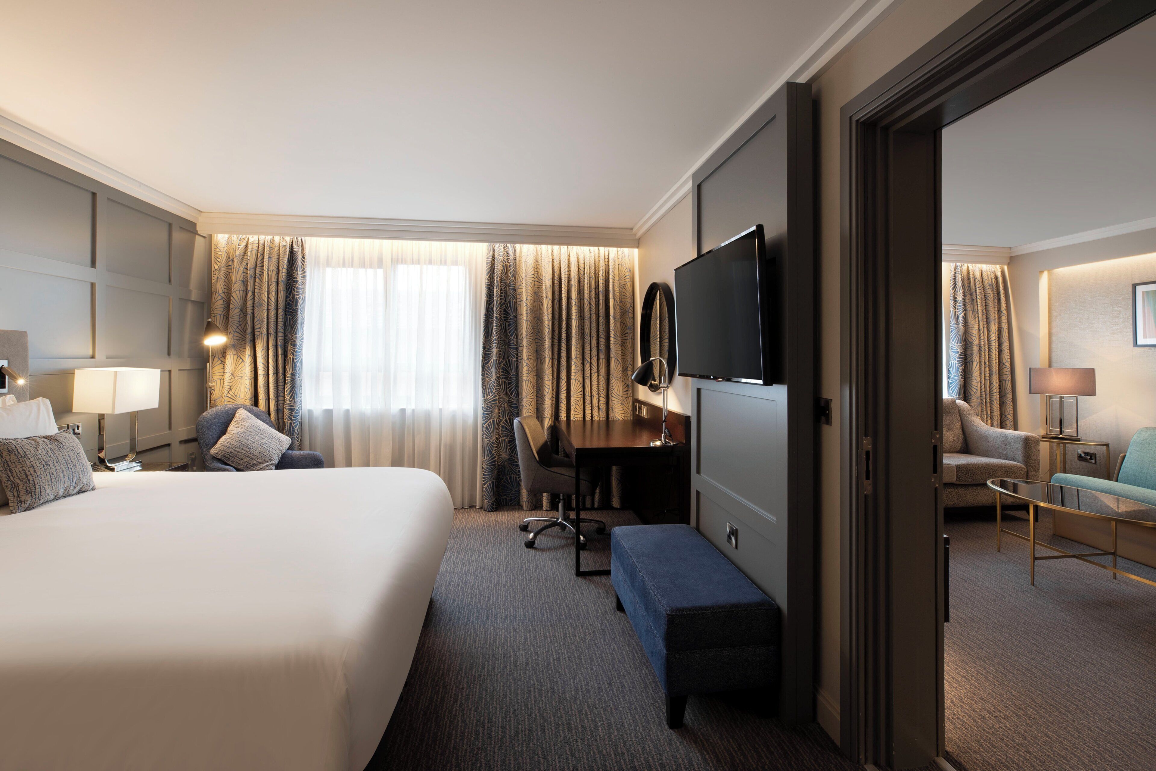 A hotel room with a large bed, a desk, a TV, and a seating area with a sofa and armchairs.