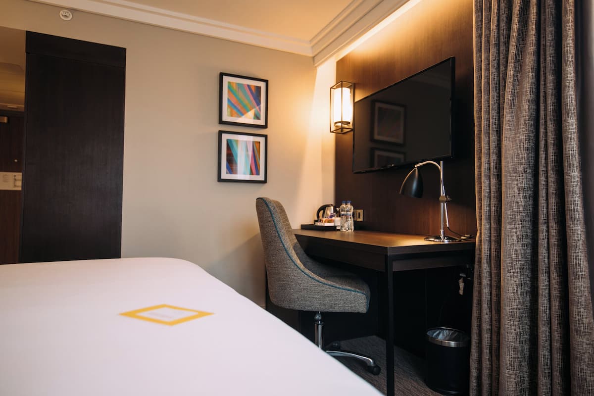 A hotel room with a bed, a desk, a chair, a desk lamp, a trash can, and two framed pictures on the wall.