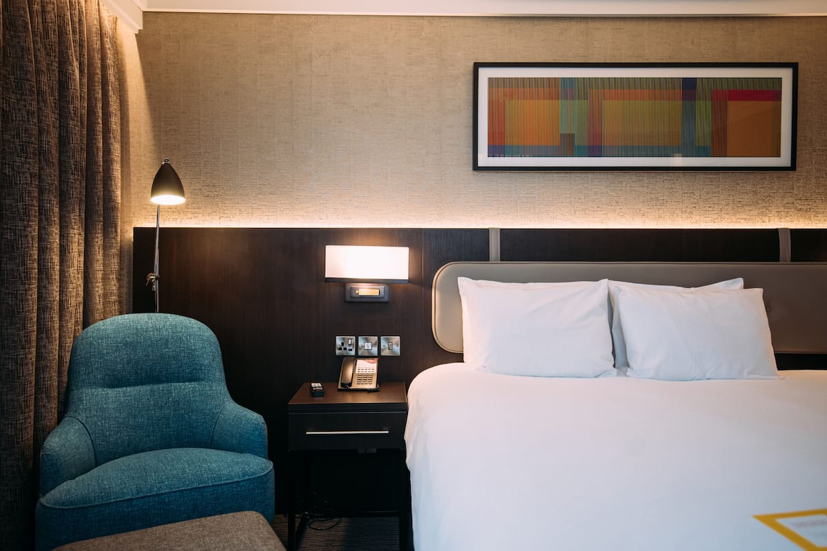 Ett hotellrum med en säng, en stol, ett nattduksbord med en lampa och en inramad tavla på väggen.
