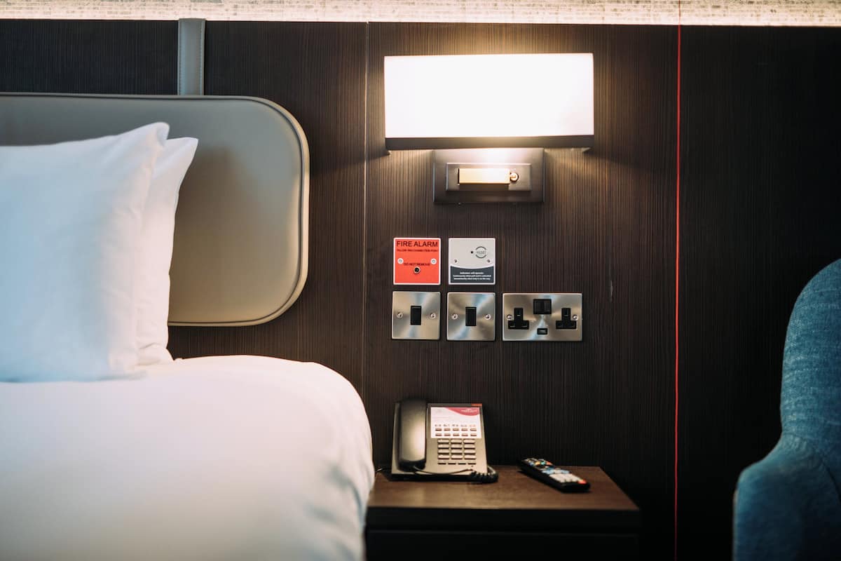 A hotel room with a bed, a bedside table with a phone and remote control, a wall-mounted light, and a fire alarm panel.