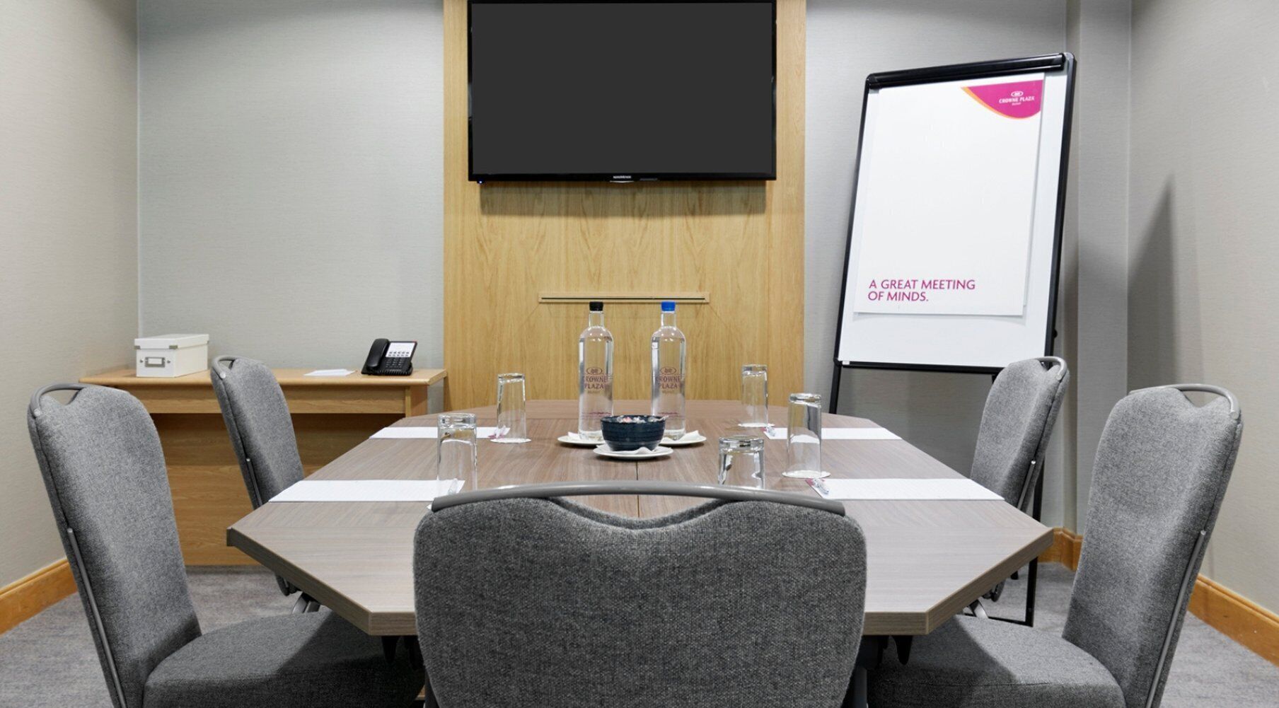A conference room with a long table, six grey chairs, a whiteboard with 'A GREAT MEETING OF MINDS' written on it, a projector screen, and a small table with a phone and a water bottle.