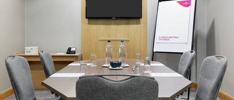 A conference room with a long table, six grey chairs, a whiteboard with 'A GREAT MEETING OF MINDS' written on it, a projector screen, and a small table with a phone and a water bottle.