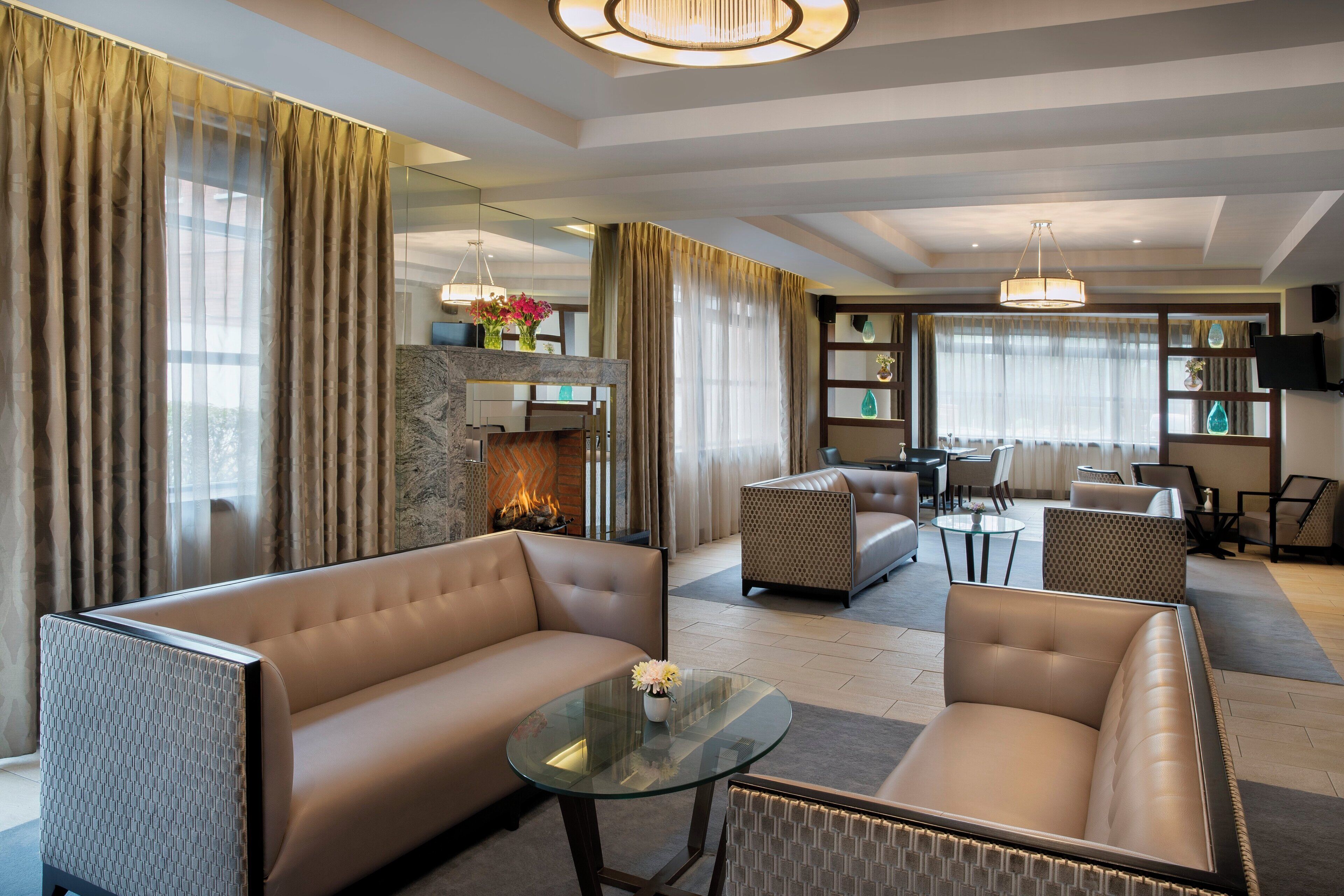 A modern living room with a sofa, armchair, and a glass coffee table. There is a fireplace with a fire, a television mounted on the wall, and a dining area with chairs and a table.