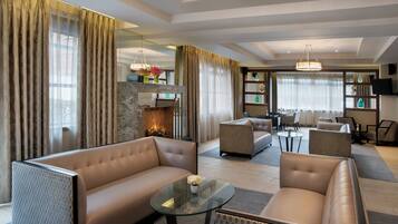 A modern living room with a sofa, armchair, and a glass coffee table. There is a fireplace with a fire, a television mounted on the wall, and a dining area with chairs and a table.