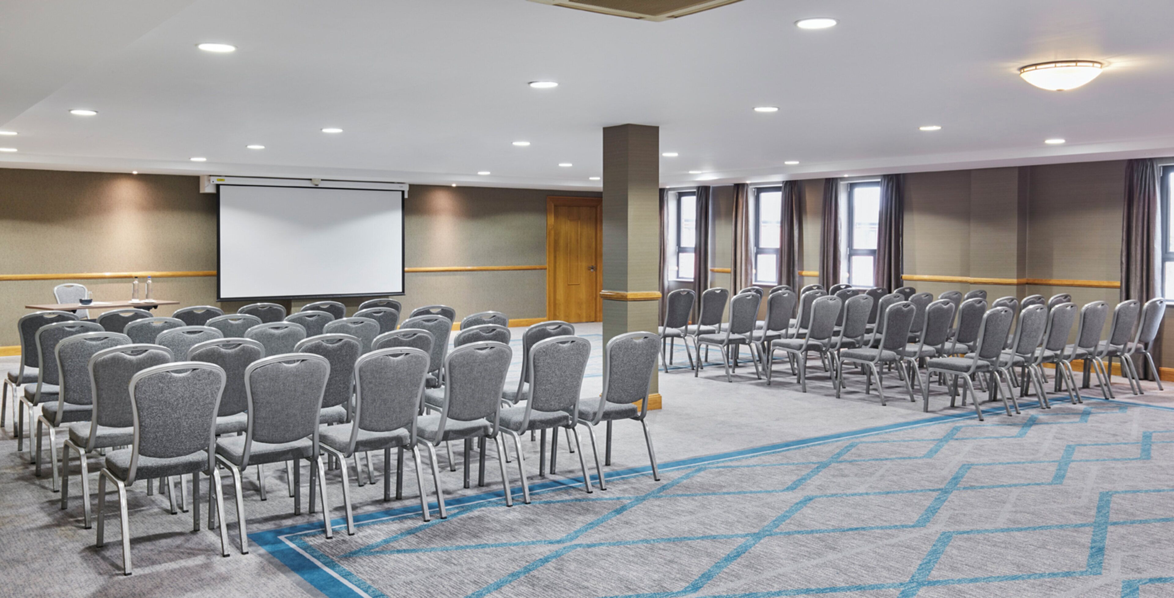 Konferenssihuone, jossa on riveissä tuoleja, projektorikangas ja matto lattialla.