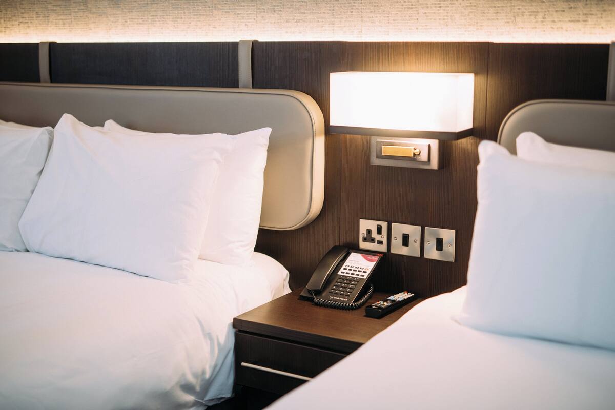 A hotel room with two beds, a bedside table with a phone and remote control, and a wall-mounted light fixture.