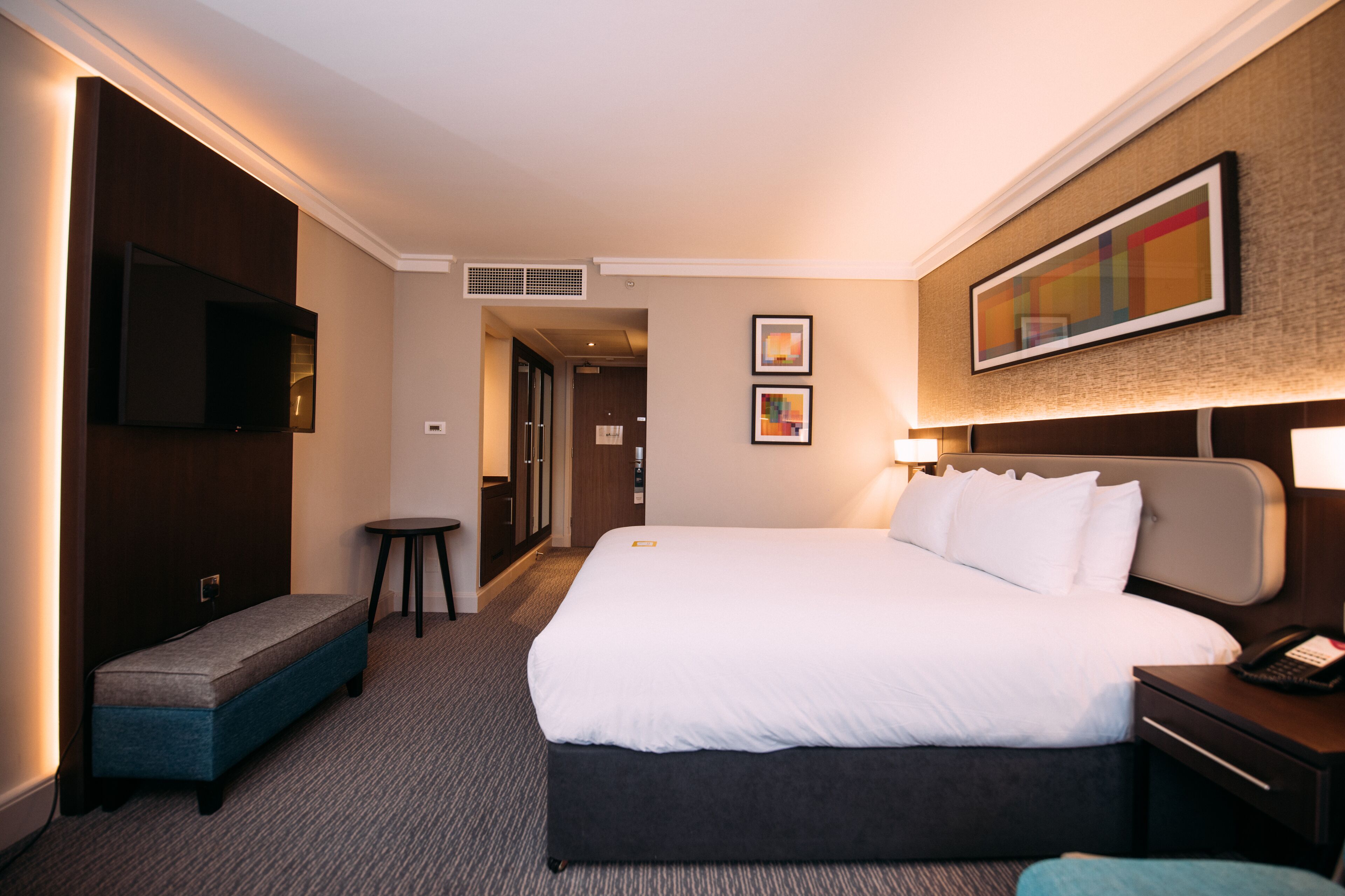 A hotel room with a large bed, a bedside table with a phone, a bench, a TV mounted on the wall, and two framed pictures on the wall.