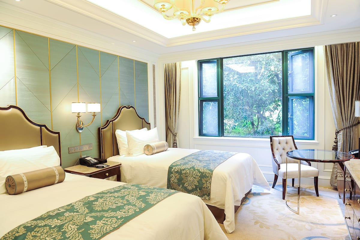 A hotel room with two beds, a sitting area with a table, and a large window offering a view of greenery.