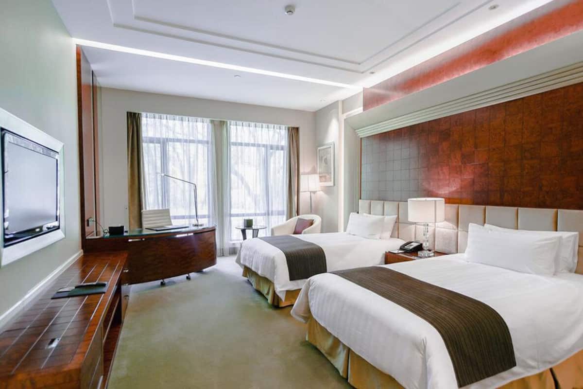 A hotel room with two beds, a desk, a TV, and a large window.