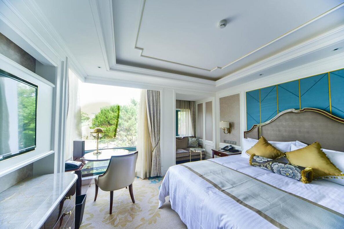 A hotel room with a large bed, a desk, and a view of greenery outside.