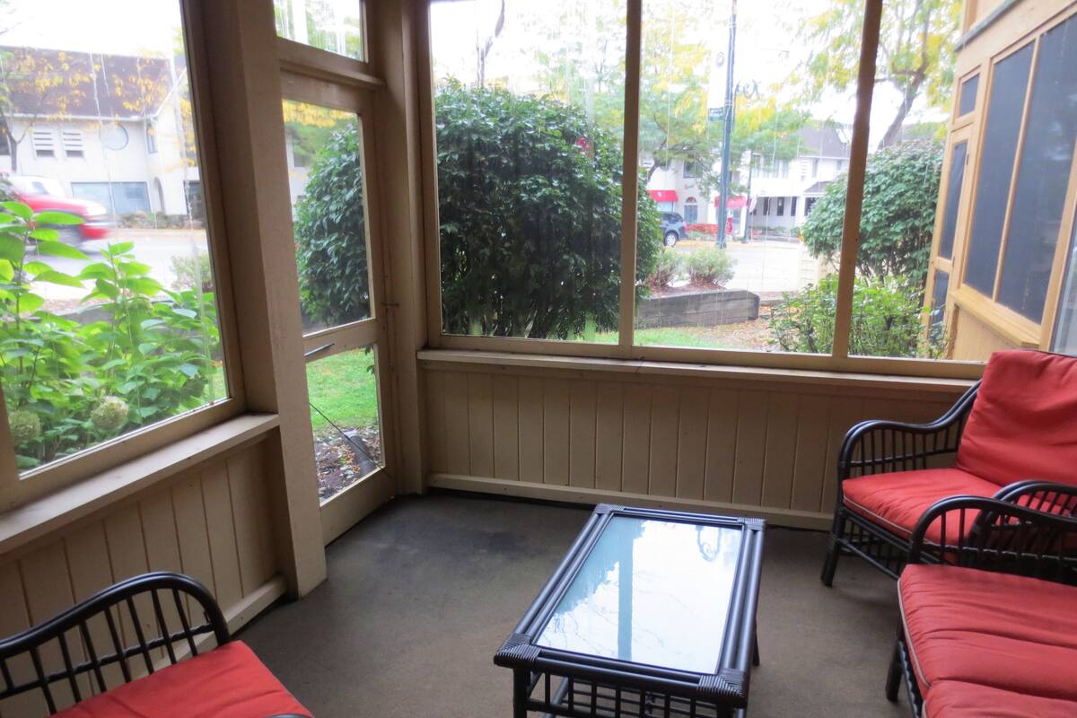 A covered porch with a glass table and two chairs, a view of a garden, and a house in the background.