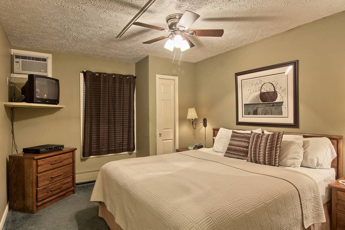 A bedroom with a bed, a ceiling fan, a television on a shelf, a door, a framed picture, and a nightstand.