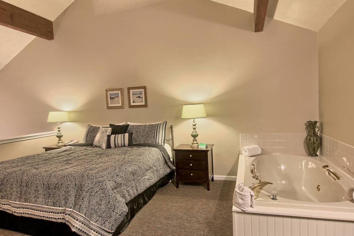 A bedroom with a large bed, two bedside tables with lamps, a small drawer, a large bathtub, and two framed pictures on the wall.