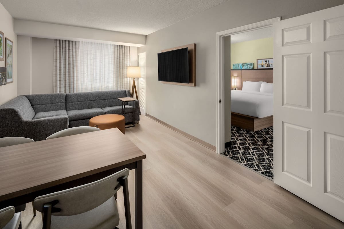 A hotel room with a gray sofa, a small round table, a wooden desk, a bed with white bedding, and a TV mounted on the wall.