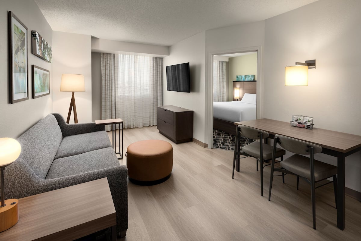 A modern hotel room with a sofa, ottoman, dining table, chairs, a TV, and a bed.