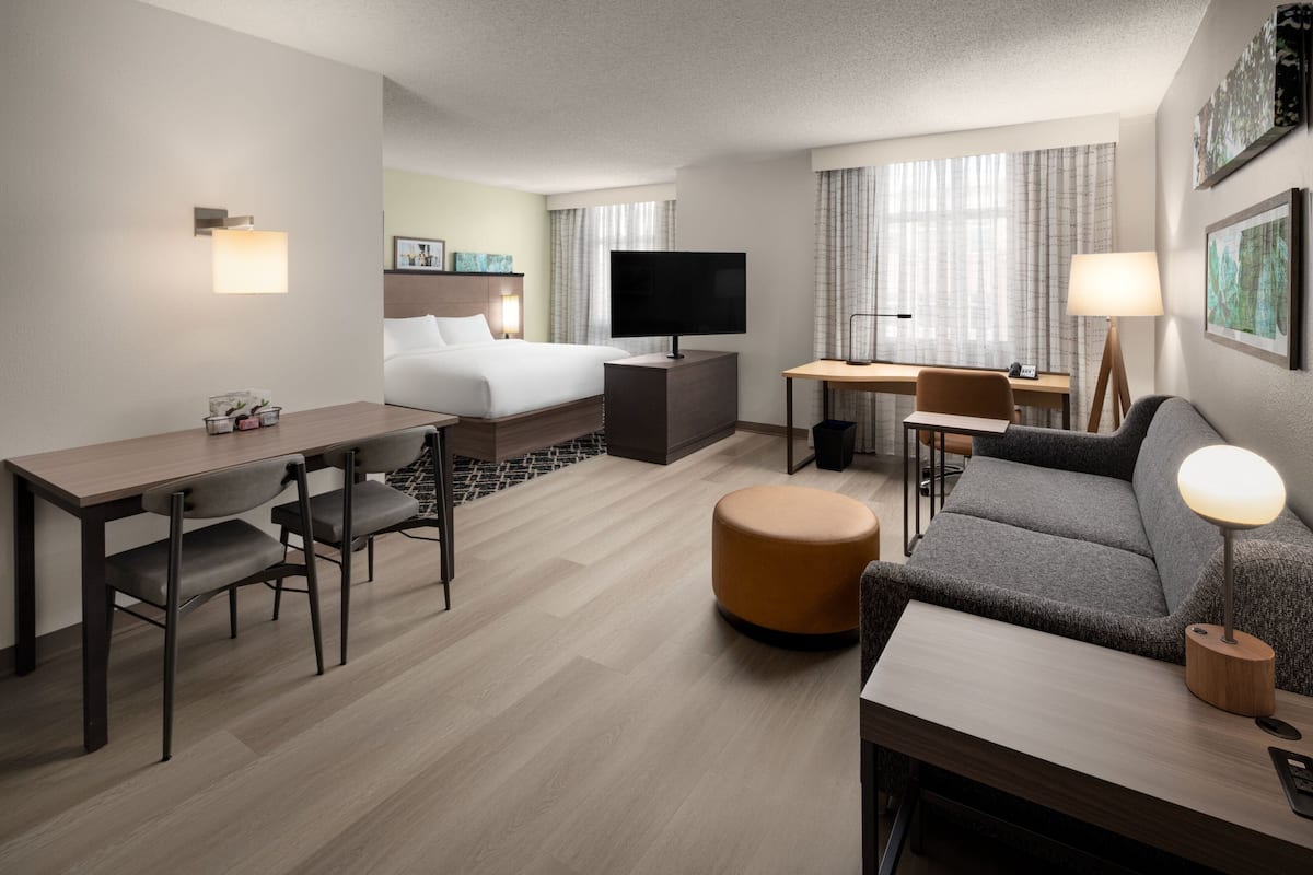 A hotel room with a bed, desk, sofa, and ottoman. There is a television, a lamp, and a picture on the wall.