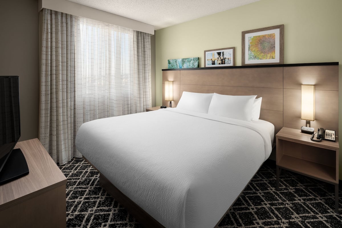 A hotel room with a large bed, two bedside tables with lamps, a TV on a stand, and a window with curtains.