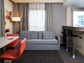 A compact hotel room with a grey sofa, a small table with a chair, a bar area, and a window with curtains. - Aparthotel Adagio La Défense Esplanade (Puteaux)