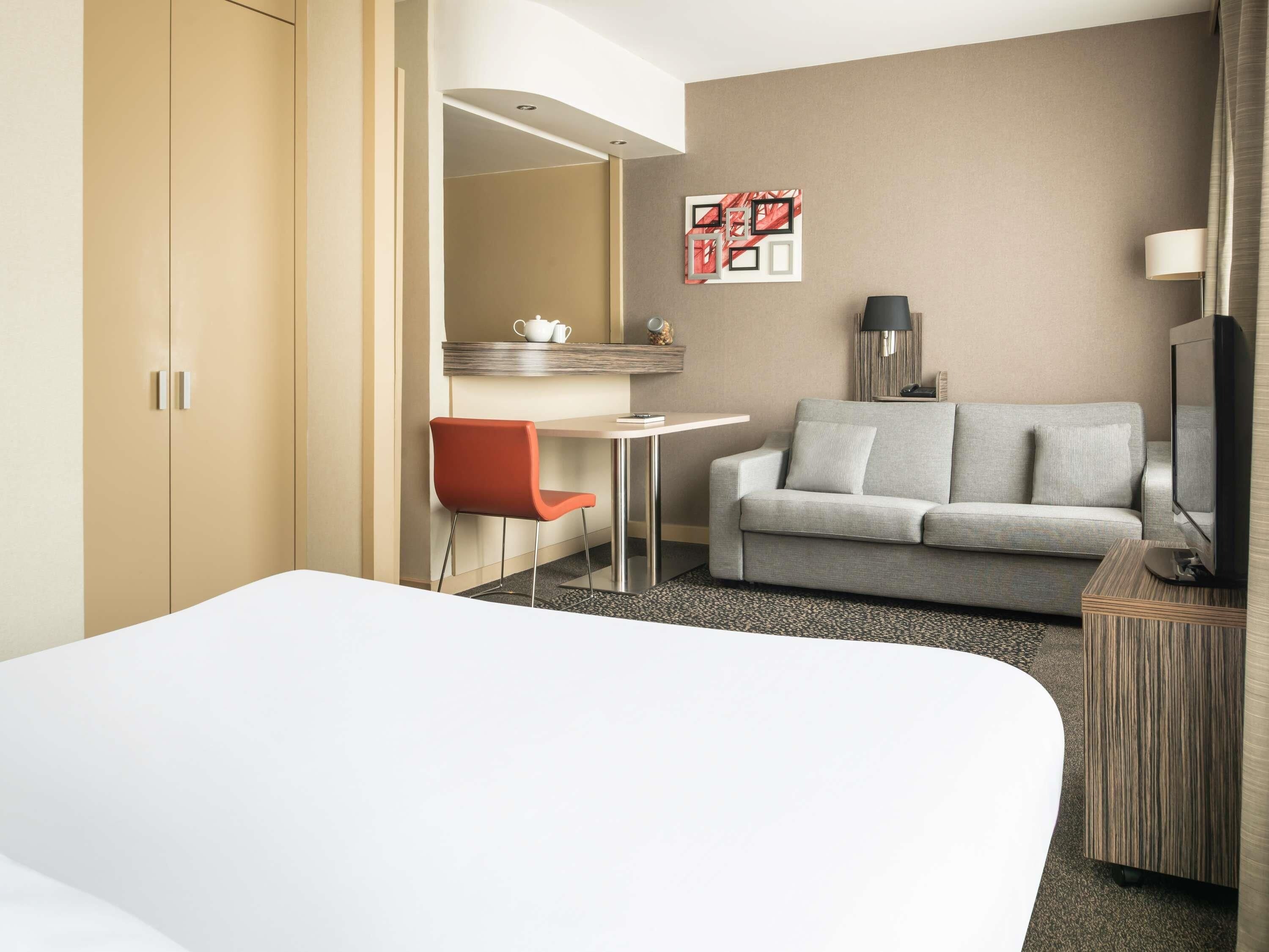 Une chambre d’hôtel avec un lit, un canapé gris, un fauteuil rouge, une petite table, un téléviseur et une œuvre d’art accrochée au mur.