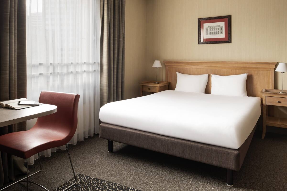 Une chambre d’hôtel avec un lit, deux tables de chevet, un bureau avec une chaise, une chaise rouge et une fenêtre avec des rideaux.