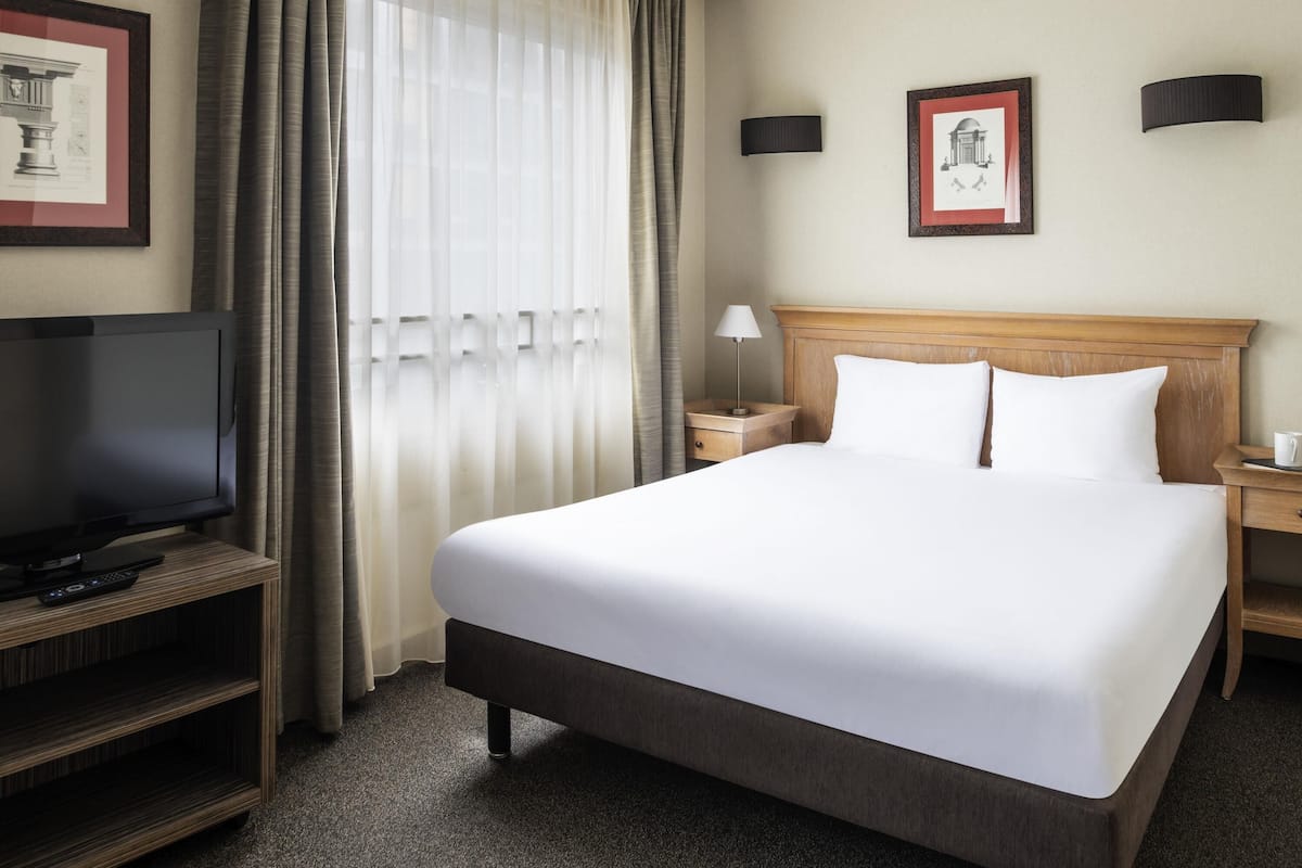 Une chambre d’hôtel avec un grand lit, un téléviseur posé sur un meuble en bois, une table de chevet et une fenêtre avec des rideaux.