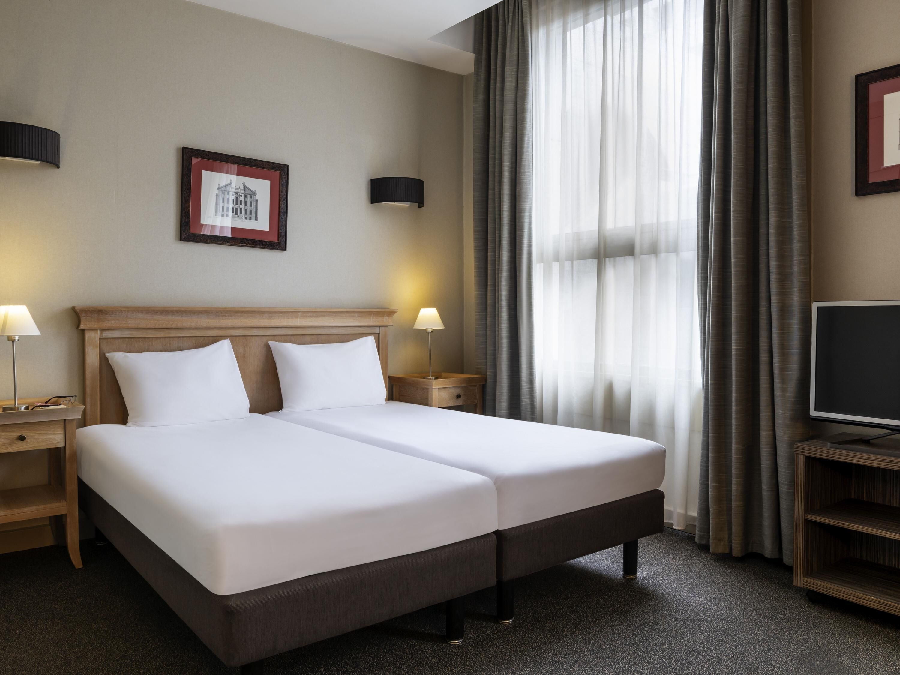 Habitación de hotel con cama doble, dos mesitas de noche con lámparas, un televisor sobre un mueble bajo, un cuadro en la pared y cortinas de encaje.