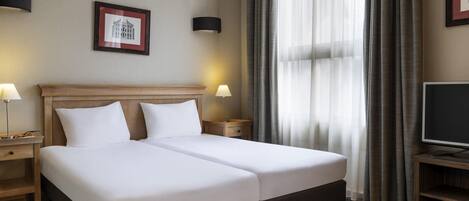 A hotel room with a double bed, two bedside tables with lamps, a TV on a stand, a framed picture on the wall, and sheer curtains.