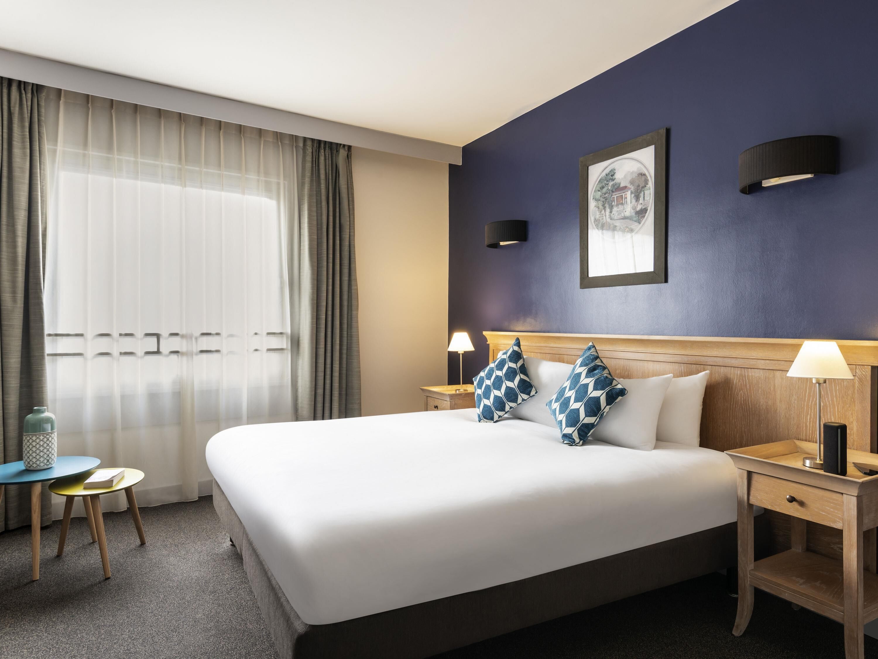 Habitación de hotel con una cama grande, dos mesitas de noche, una mesita auxiliar pequeña, una ventana con cortinas translúcidas y una pared de acento azul oscuro.