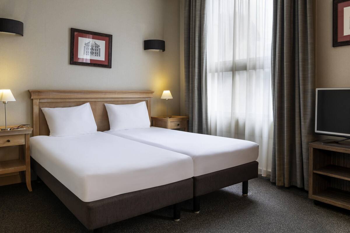 Une chambre d’hôtel avec un lit double, deux tables de chevet avec des lampes, un téléviseur posé sur un meuble, un tableau encadré au mur et des rideaux légers.