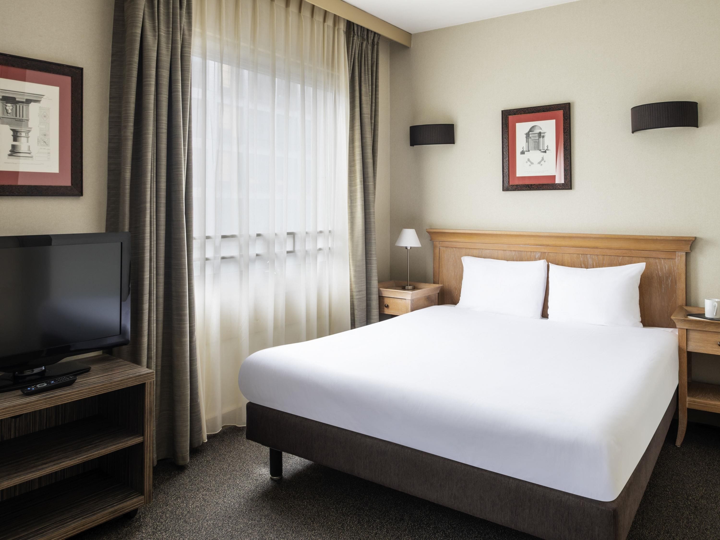A hotel room with a large bed, a television on a wooden stand, a nightstand, and a window with curtains.
