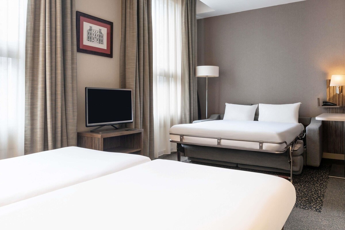 A hotel room with two beds, a flat-screen TV on a wooden stand, a bedside table, a lamp, and a framed picture on the wall.