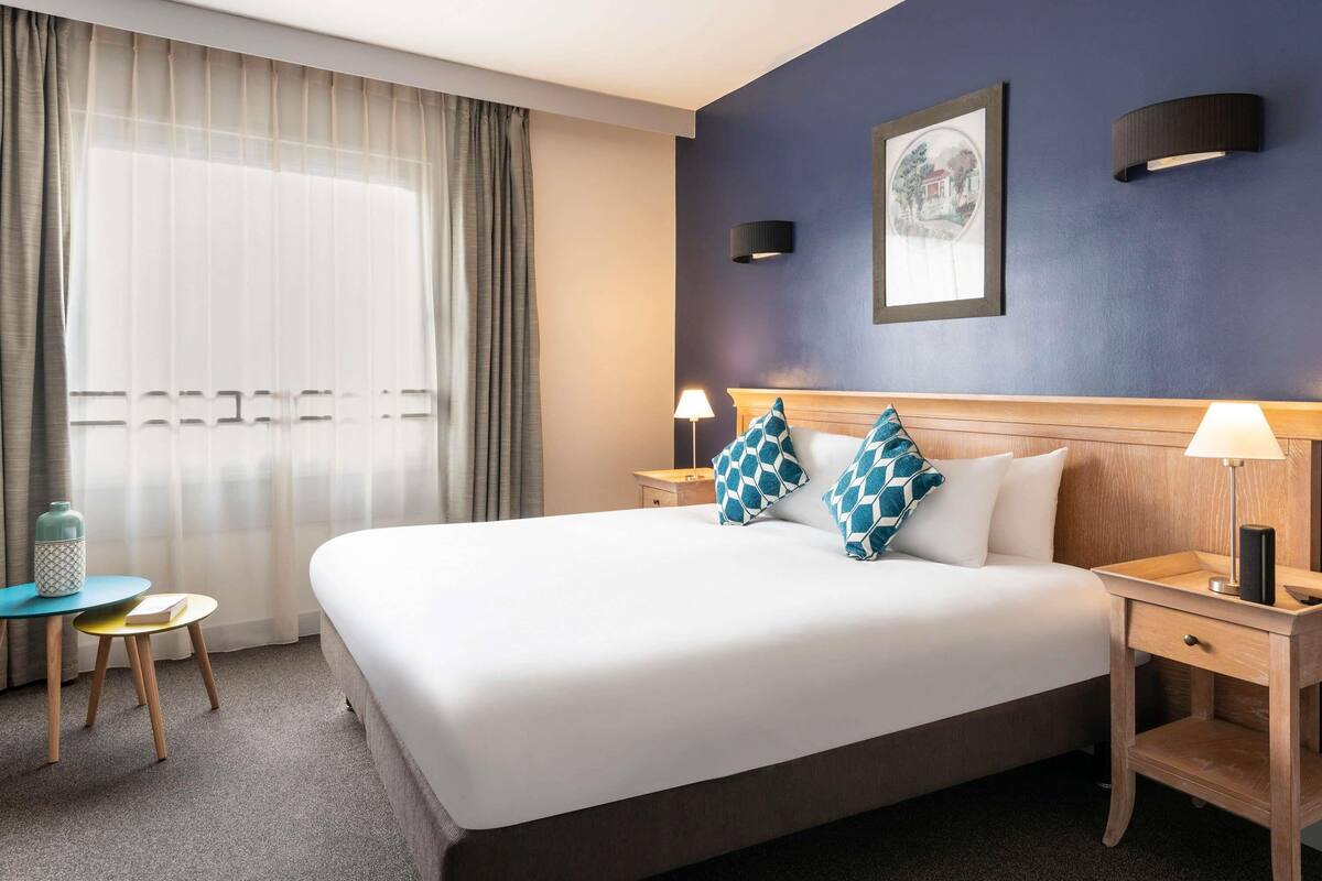 Une chambre d’hôtel avec un grand lit, deux tables de chevet avec des lampes, une petite table ronde avec un livre, et une fenêtre avec des rideaux légers.