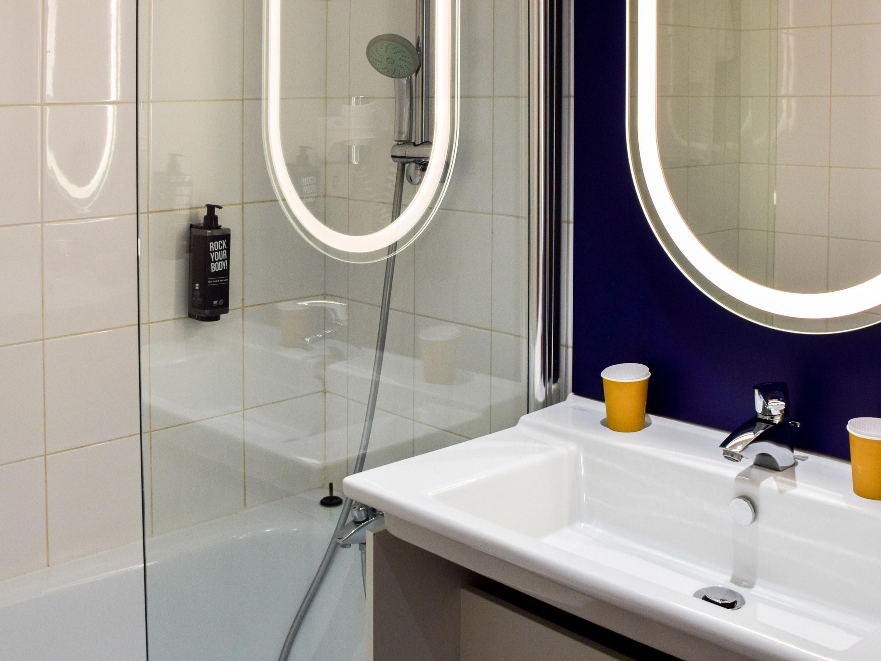 Un baño con ducha de vidrio, un lavabo blanco con dos llaves y un espejo con luz.