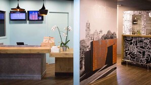 A reception area with a wooden desk, two pendant lights, a wall-mounted TV, and a mural with abstract designs. - ibis Rodez Centre Cathédrale (Rodez)