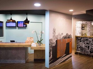 Zona de recepció amb un taulell de fusta, dues làmpades penjants, un televisor fixat a la paret i un mural amb dissenys abstractes.