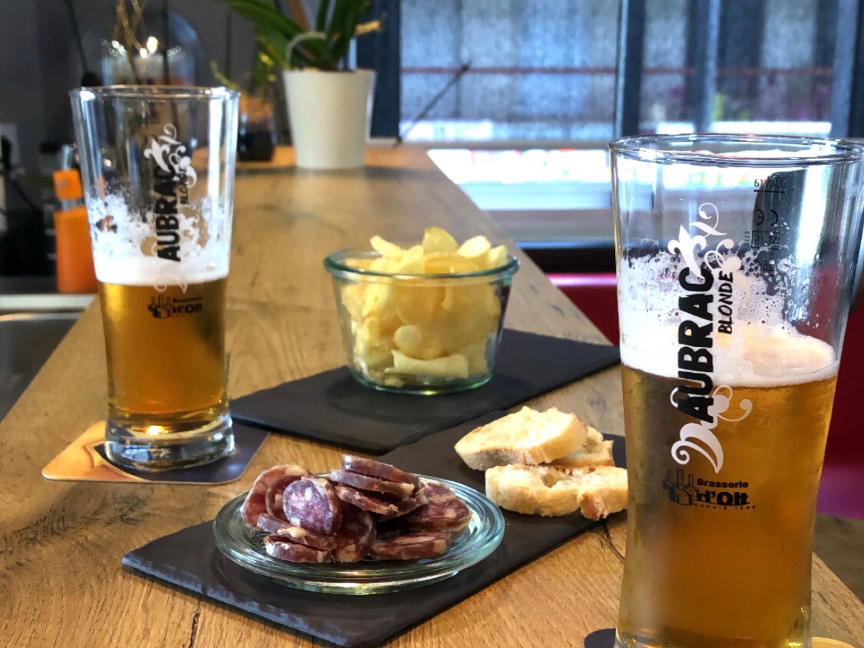 Dos vasos de cerveza sobre una mesa de madera con un bol de patatas fritas y un plato de embutidos.